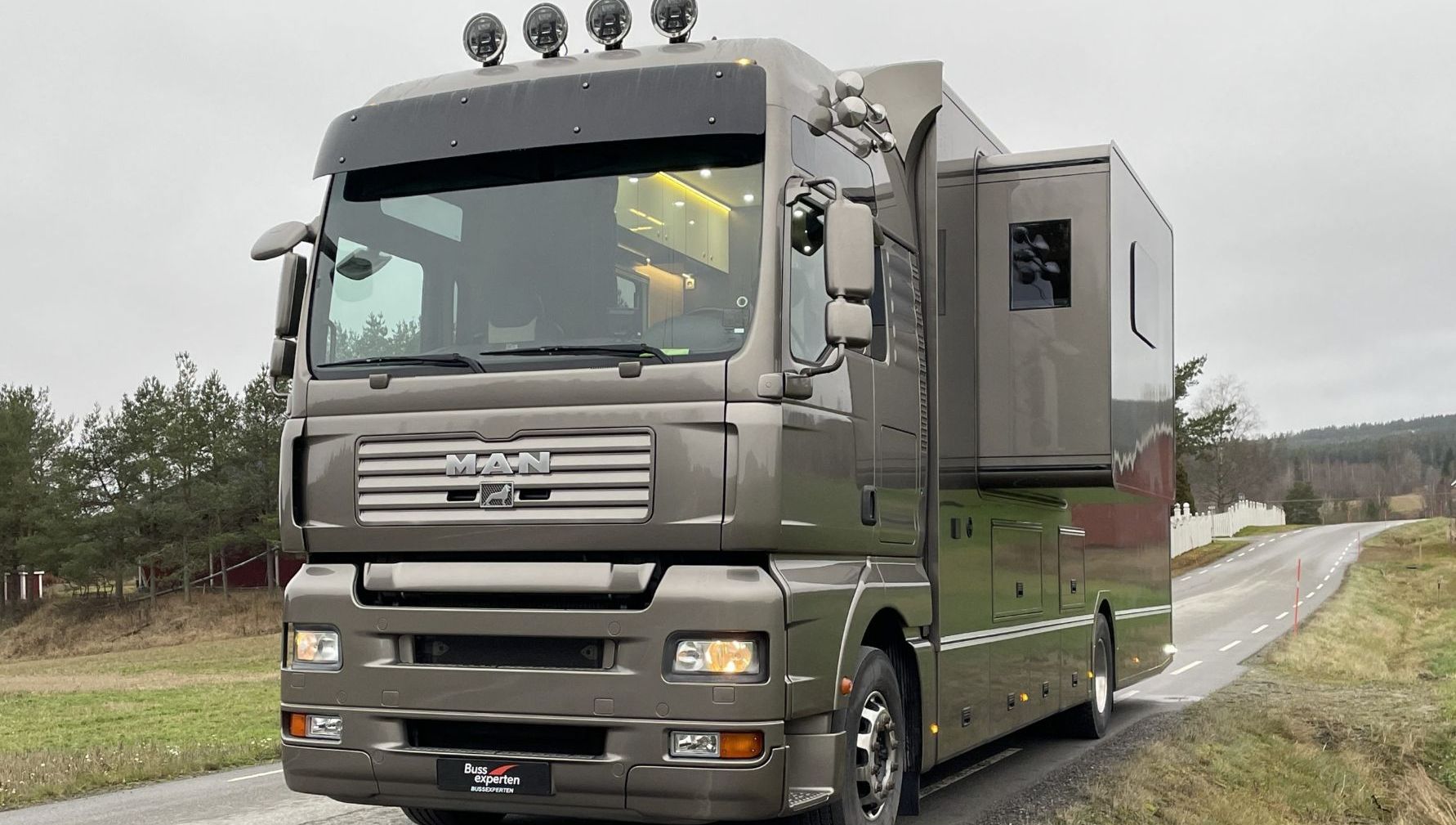 Grå lastebilbasert bobil med utvidet sidevegg og taklamper, parkert langs landevei.