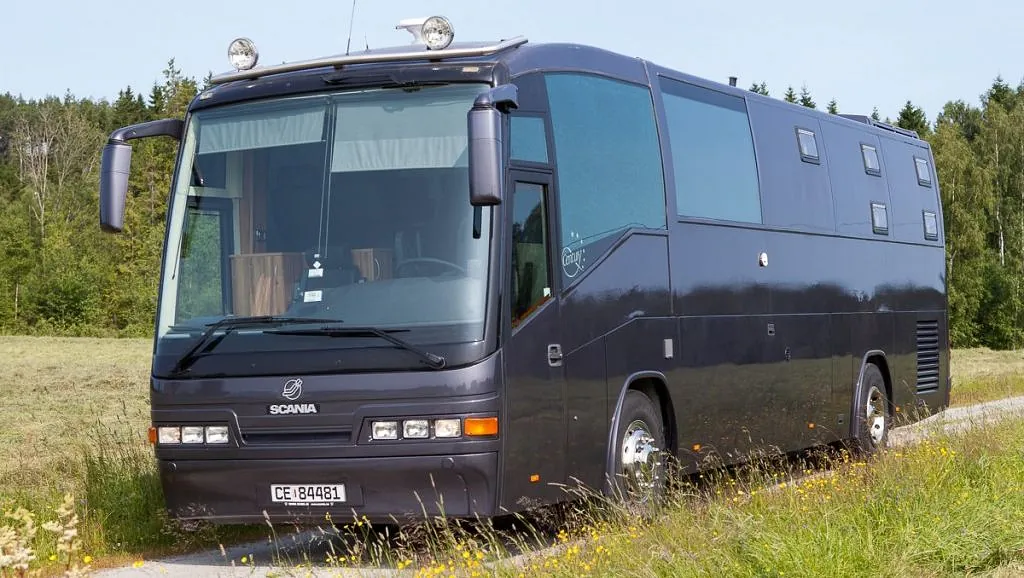 En mørk Scania-buss parkert i en eng med tørket gress, mot en klar himmel og skog i bakgrunnen.