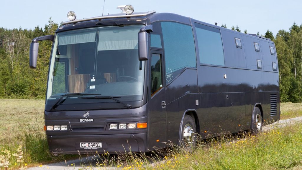 En mørk Scania-buss parkert i en eng med tørket gress, mot en klar himmel og skog i bakgrunnen.