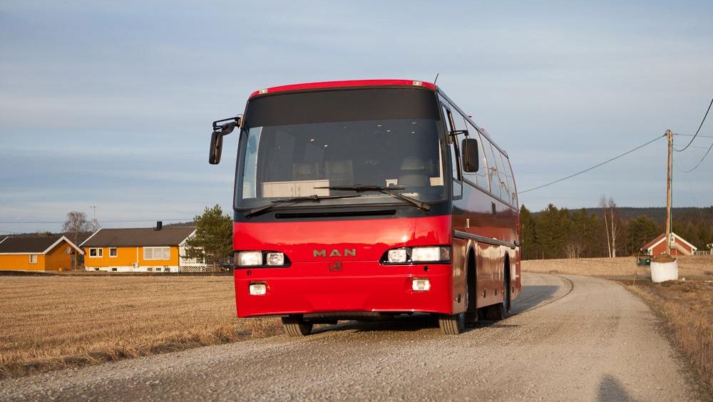 Rød MAN turbuss på landevei med hus i bakgrunnen