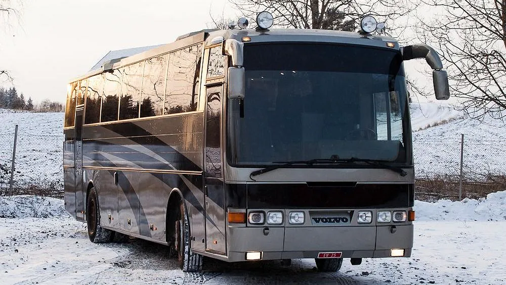 Fronten av en grå og svart Volvo buss parkert på en snødekket vei med trær i bakgrunnen