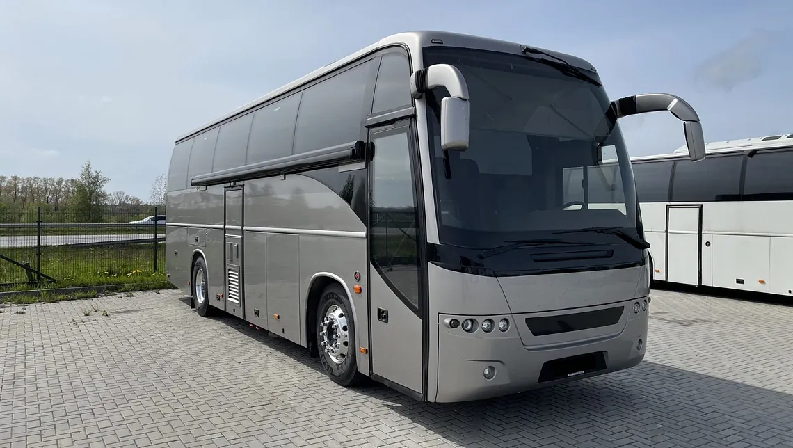 Sølvfarget turbuss sett forfra fra skrå vinkel, med store sidespeil og tonede vinduer, parkert på steinlagt område.