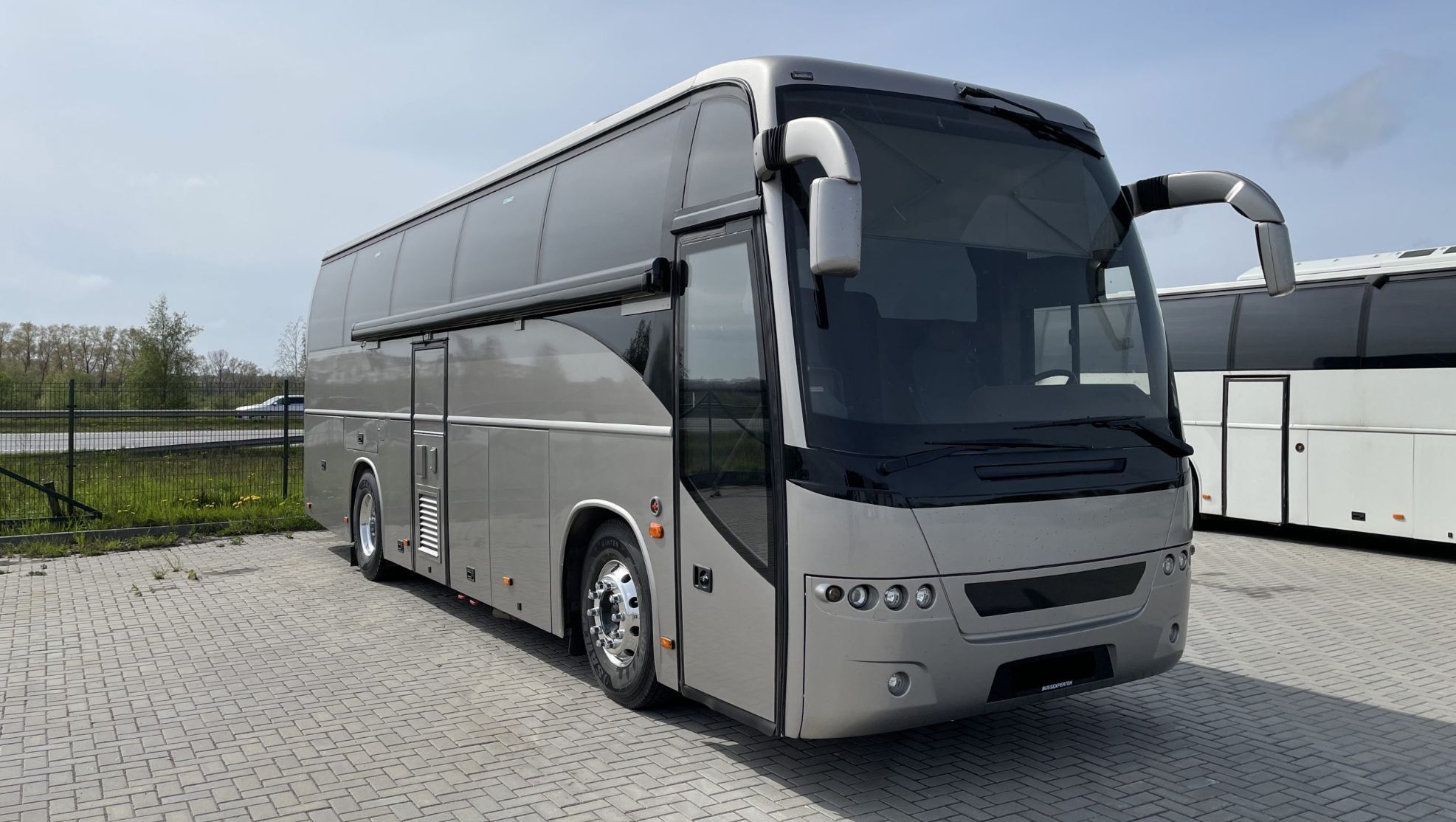 Sølvfarget turbuss sett forfra fra skrå vinkel, med store sidespeil og tonede vinduer, parkert på steinlagt område.