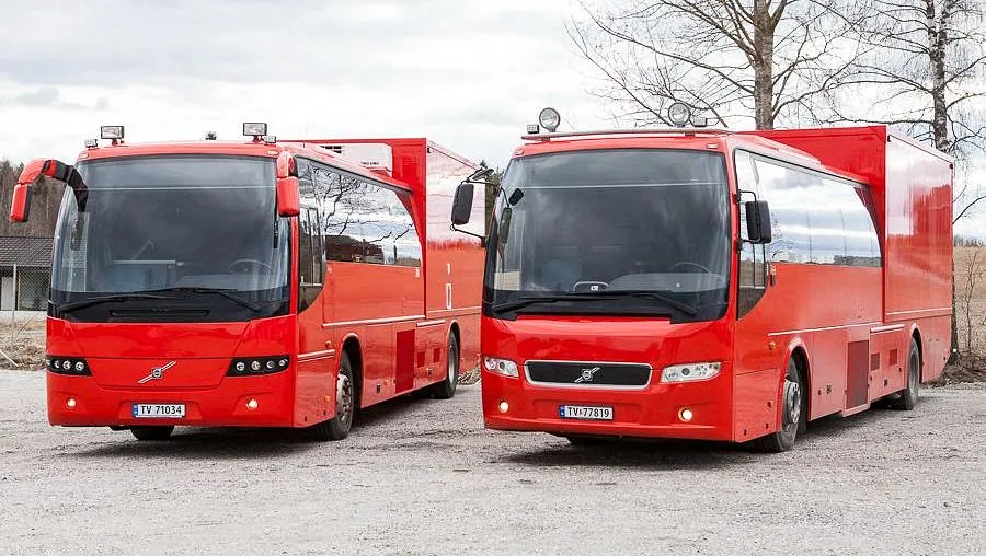 To røde Volvo-busser med påbygg parkert utendørs ved åker og trær.
