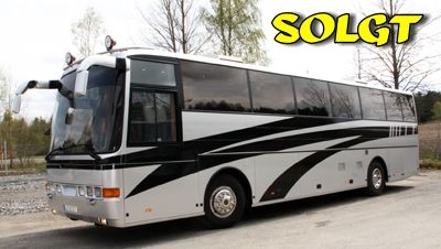 Sølv og sort turbuss med "SOLGT"-tekst øverst, parkert ved grusvei og skog.