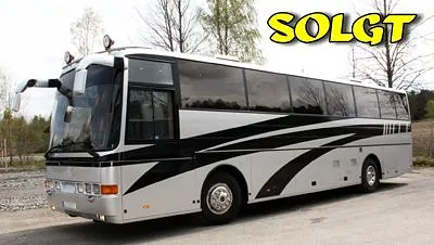 Sølv og sort turbuss med "SOLGT"-tekst øverst, parkert ved grusvei og skog.