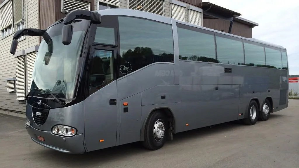 Grå turbuss med store vinduer parkert foran en bygning med liggende kledning.