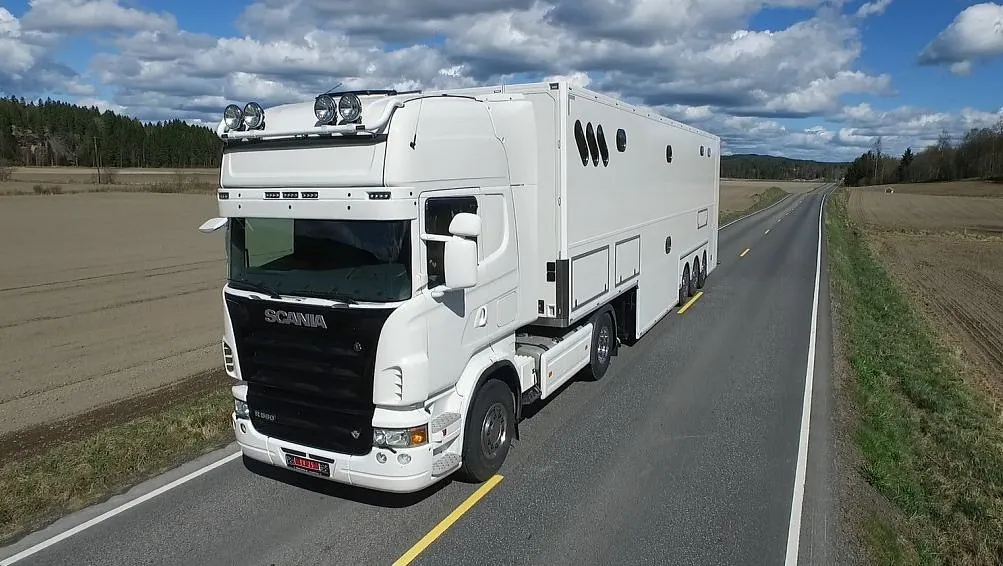 Hvit Scania lastebil med stor hvit tilhenger, kjøres på landevei i åpent landskap.