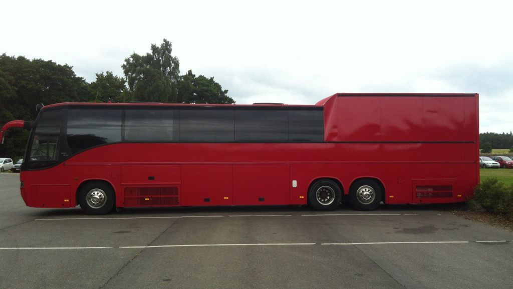 Sidebilde av lang rød turbuss med ekstra høyt lasterom bakerst, parkert ved grøntområde.