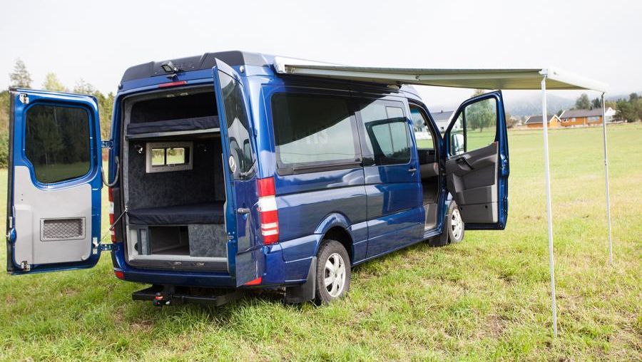 Blå campervan med åpen bakdør og markise, parkert på en gresslette.