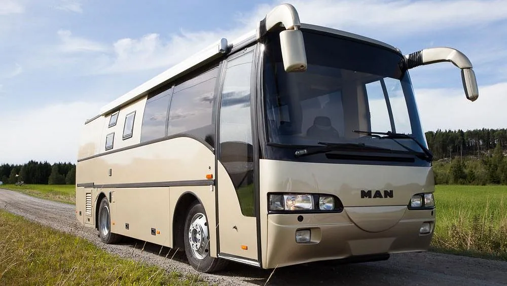 Beige MAN turbuss med sotede vinduer parkert på grusvei i landlig landskap.