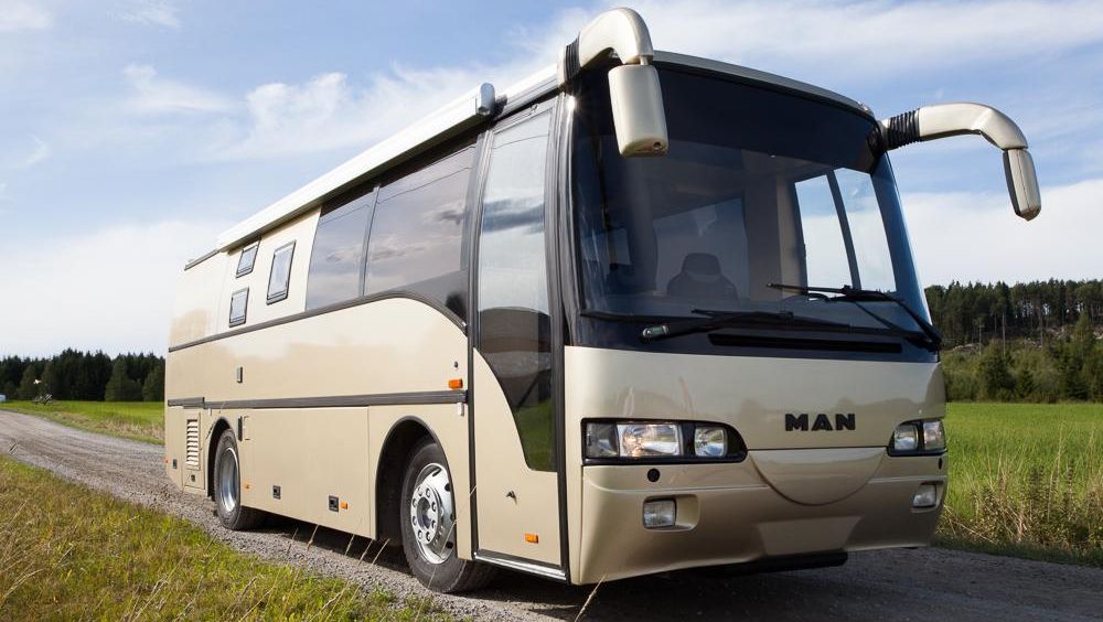 Beige MAN turbuss med sotede vinduer parkert på grusvei i landlig landskap.