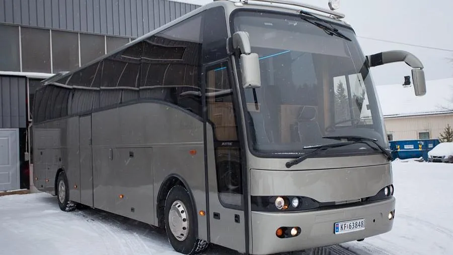 Fronten av en grå buss med lys på taket, parkert i snøen foran en grå bygning.