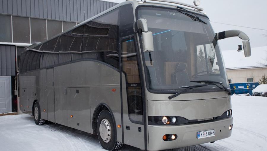 Fronten av en grå buss med lys på taket, parkert i snøen foran en grå bygning.