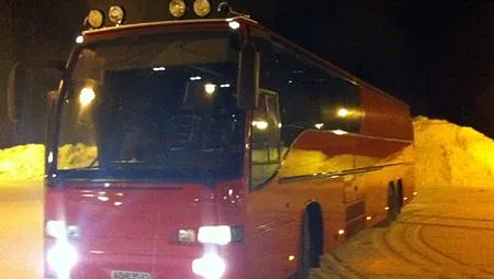 Rød turbuss med frontlykter påslått, parkert ute på snødekt plass om natten.