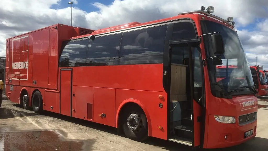 Rød buss med påbygg og Veidekke logo står med åpen dør på parkeringsplass