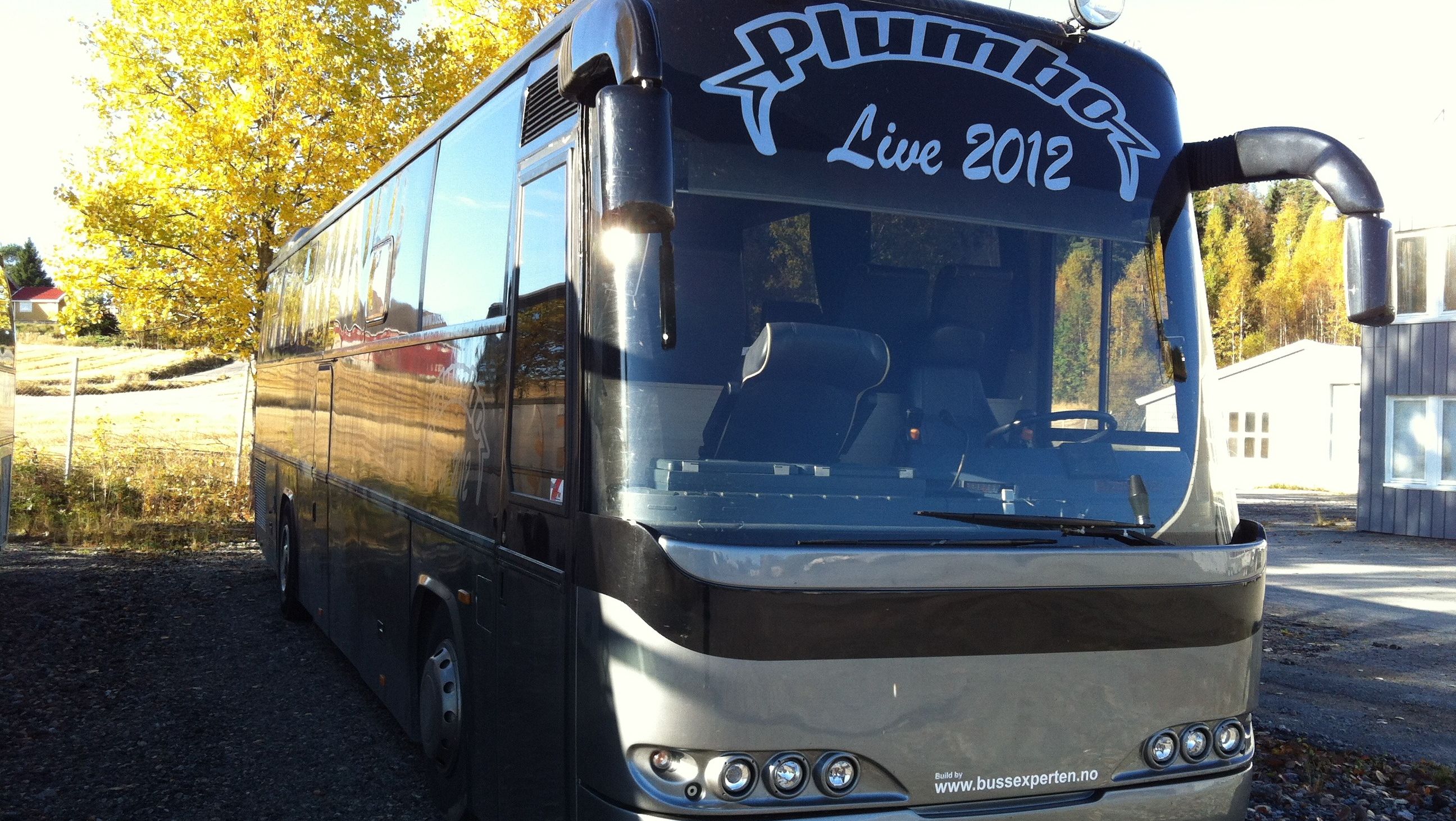 Grå turbuss parkert utendørs i høstlandskap, med Plumbo logo og "Live 2012" på fronten.