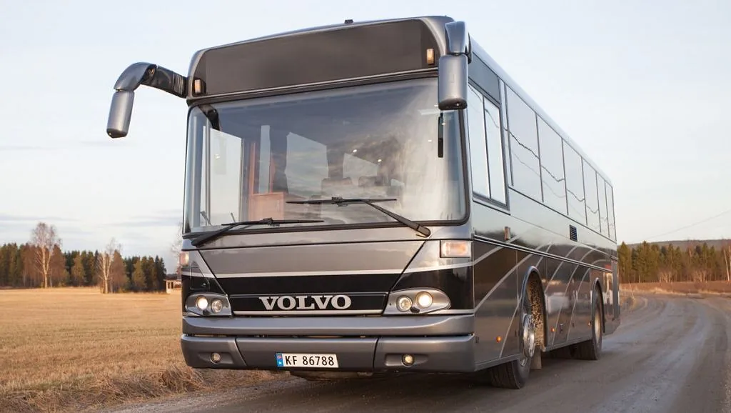 Grå og svart turbuss sett fra front kjører på grusvei i landlige omgivelser