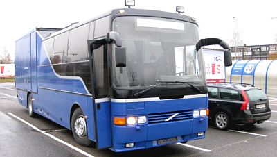 Blå Volvo turbuss med ekstra bakdel og store frontvinduer, parkert ved butikkområde.