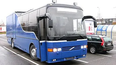 Blå Volvo turbuss med ekstra bakdel og store frontvinduer, parkert ved butikkområde.