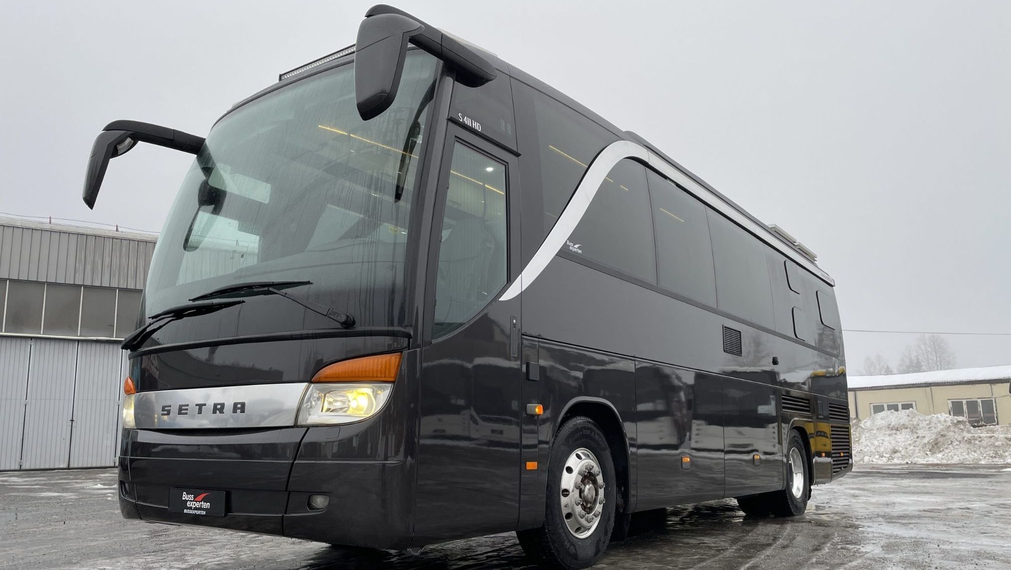 Svart turbuss med store vinduer og kromdetaljer foran, parkert på islagt underlag ved bygg.