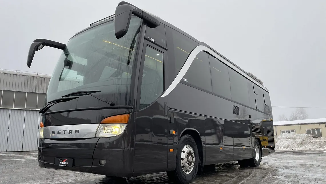 Svart turbuss med store vinduer og kromdetaljer foran, parkert på islagt underlag ved bygg.